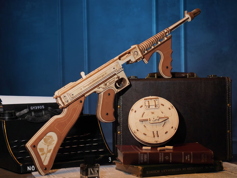 A wooden model of a submachine gun with a rose engraving on the stock, displayed on a table next to a vintage typewriter, a black briefcase with a clock face, and books against a dark blue paneled background.